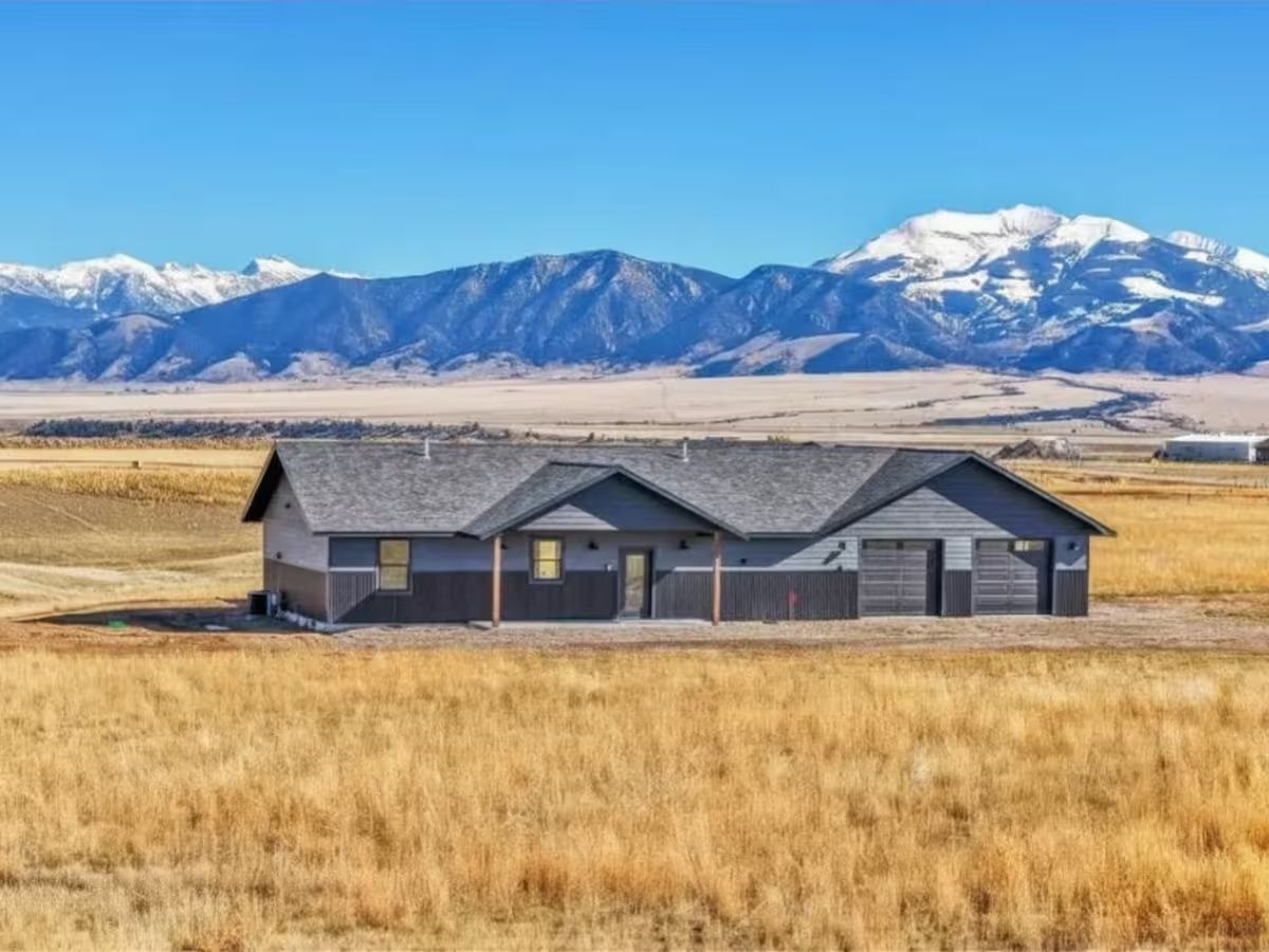 A single-storey home with dark siding on open grassland outside Ennis, snow-capped mountains across the valley behind it.