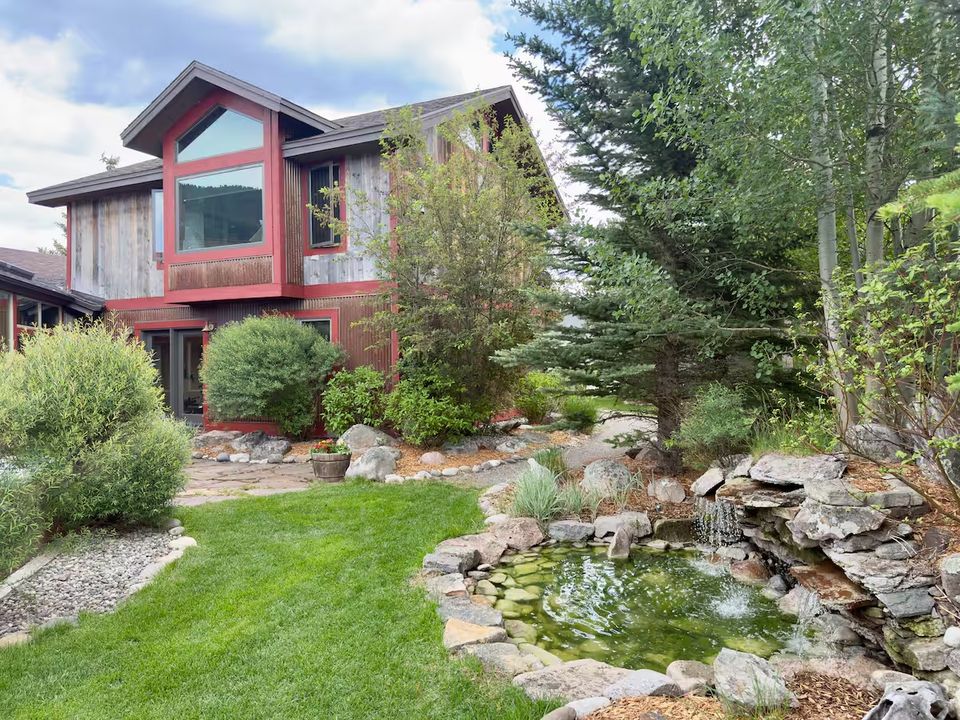 The back of the house in summer: weathered barn-wood siding with red trim, tall gabled windows, and a stone-edged pond with a small waterfall in the garden.