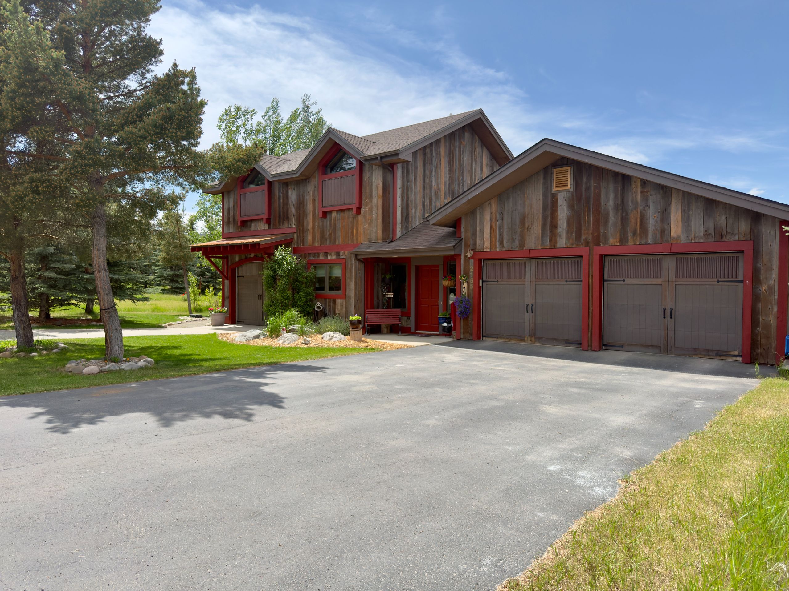 The Yellowtail house in summer: two storeys clad in weathered barn wood with red-painted window trim, a red front door and a three-bay garage, with pines and mown lawn along the driveway.